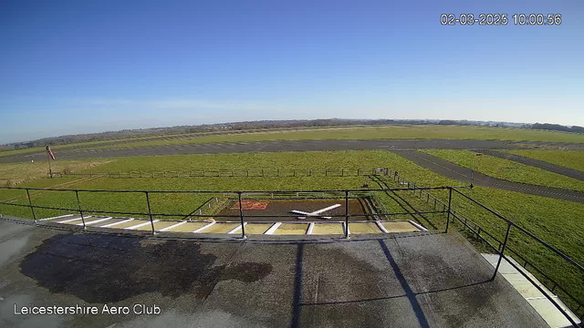 A clear blue sky stretches overhead, with no clouds visible. Below is a large expanse of green grass, representing a field or runway. There is a circular helipad marked with a white cross in the center, surrounded by a concrete area. On the left side, a small wind indicator, resembling a flag, is mounted on a pole. The background features a winding path and additional grassy areas, with a fence line demarcating the boundaries. The scene conveys a tranquil outdoor environment, likely part of an aerodrome or airfield.