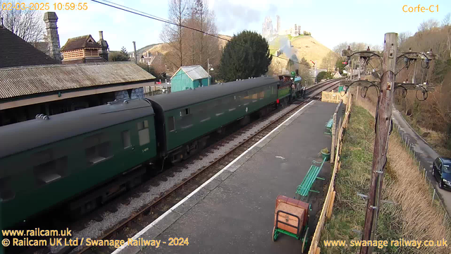 A green vintage steam train is passing through a railway station. The train has several green carriages and is approaching from the left side of the image. In the background, there is a hill with a castle at the top, partially obscured by smoke from the train. To the right, there is a wooden utility pole with wires and a few benches along the platform. The setting is bright and sunny, with trees and buildings visible on the station platform.