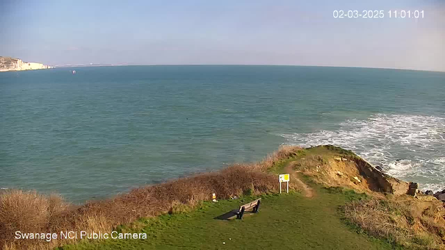 A coastal scene showing a vast blue sea under a clear sky. To the right, cliffs rise from the water, and patches of greenery are visible on the shore. In the foreground, there is a wooden bench facing the ocean, situated on grassy land bordered by shrubs. A caution sign can be seen on the path leading towards the cliff's edge.