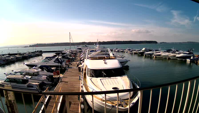 A marina with multiple boats docked along a wooden pier. In the foreground, a large white yacht is visible. Surrounding it are smaller boats, some covered and some uncovered. The water is calm and reflects the blue sky, which has a few wispy clouds. A distant shoreline is faintly visible. The scene conveys a tranquil, sunny day at the docks.