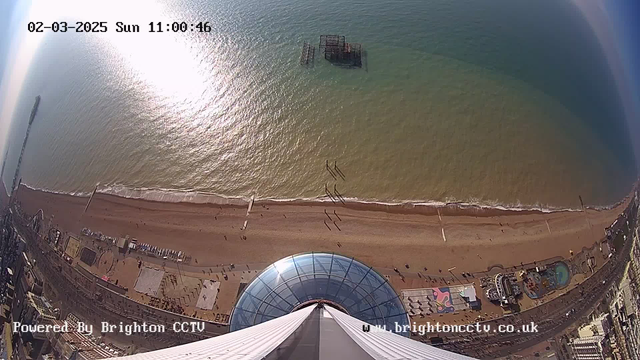 A view from a high vantage point overlooking a sandy beach and the sea. The water reflects sunlight, creating a shimmering effect on the surface. In the distance, a pier extends into the water with a structure partially submerged. The beach is lined with people walking, and various amusement rides and attractions are visible on the shore. The sky is clear with a few clouds. The bottom part of the image shows a circular glass structure, possibly part of an observation deck. The date and time are displayed in the top left corner.