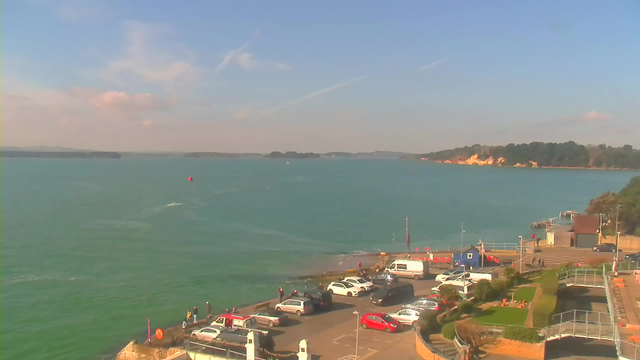 A coastal view featuring a calm turquoise sea under a mostly clear sky with a few clouds. In the foreground, there is a parking area filled with various parked cars. A few people are standing along the edge of the water, observing the surroundings. A red buoy is visible in the water, and some small boats can be seen in the distance. To the right, there is a dock area with buildings and greenery lining the shore. In the background, a rugged coastline extends, partially shrouded by trees.