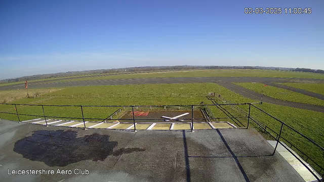 A clear blue sky with no clouds stretches overhead, and the landscape below features a large, open green field. In the distance, there is an airstrip with a dark, paved runway running horizontally across the field. A few small structures, including a windsock on a pole and a marked area for aircraft with a red outline, are visible nearby. The photo is taken from a high vantage point, likely from a building or lookout. The foreground shows a flat rooftop surface with some water marks. The scene conveys a peaceful, sunny day at an airfield, with no aircraft in sight.