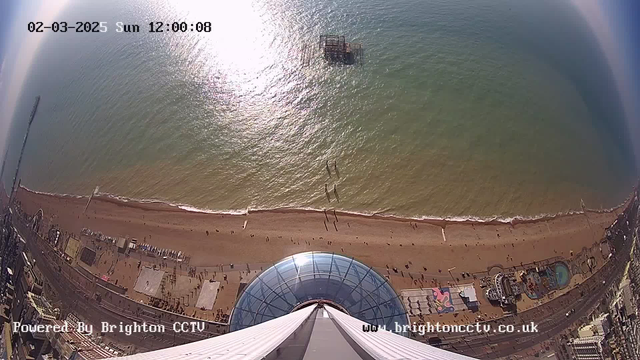 Aerial view of Brighton beach on a sunny day, with a sandy shoreline stretching along the bottom of the image. The sea, reflecting sunlight, appears to blend into a clear blue sky. People can be seen walking along the beach, and a pier extends out into the water with some structure visible above the waves. In the foreground, a large glass dome is prominent, likely a part of an observation or entertainment facility. The scene captures a lively coastal atmosphere.