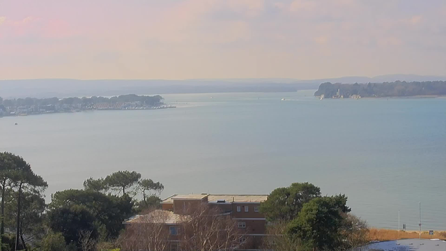 A panoramic view of a tranquil body of water, with soft blue and gray hues reflecting the sky. In the foreground, there is a multi-story building with several windows, surrounded by trees. In the distance, you can see a shoreline lined with houses and boats, leading to a gentle rise of hills. The atmosphere appears calm and serene, with a few scattered clouds in the sky.