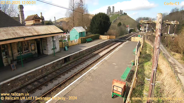 A view of Corfe Castle railway station on a sunny day. The station features a stone building with a sloped roof and large windows, along with green benches on the platform. There is a green wooden shed in the background and a sign on the ground indicating "WAY OUT." Train tracks extend down the platform, leading toward the distant cliffs where Corfe Castle sits atop a hill. Surrounding the area are trees and sparse vegetation, with a dirt path visible beside the tracks.