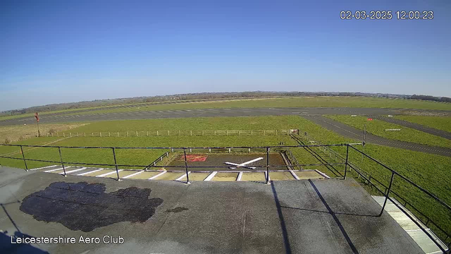 A clear blue sky fills the upper half of the image. Below, an expansive green grassy area is visible, with a section of grey pavement marked for landing. A white X marking and a red symbol can be seen on the pavement, indicating the location of a helipad. In the background, a wooden fence borders the grassy area, leading towards a distant tree line. A windsock stands on the left side, indicating the wind direction. The overall scene captures an airfield setting, with no aircraft present.