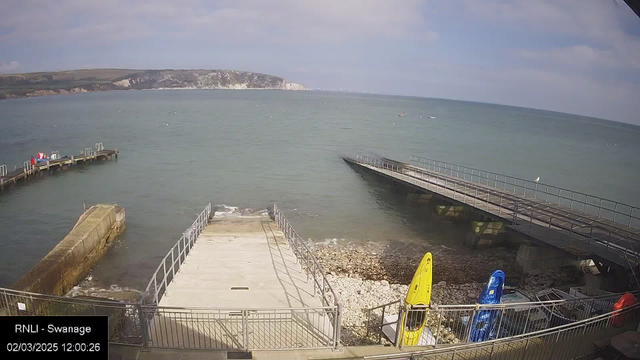 A coastal scene featuring a calm sea under a partly cloudy sky. In the foreground, there is a concrete ramp leading down to the water, bordered by a low railing. To the right, a small dock extends into the water, while a second dock is visible in the distance, both made of dark material. On the shore, several colorful kayaks are parked: a yellow one, a blue one, and a red one. The water shows some gentle ripples with a few boats visible further out. The background includes cliffs and greenery.