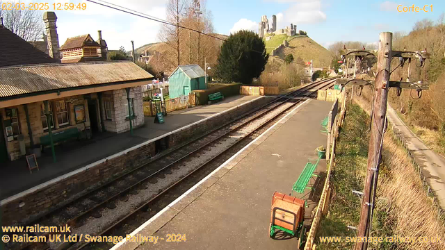 The image shows a railway station platform with tracks visible extending to the right. To the left, there is a stone building with a sloped roof and a green bench in front. A small wooden sign stands on the platform. In the background, a green shed is situated behind some benches, and beyond that, there is a picket fence. On the hillside in the distance, the ruins of a castle are prominently featured against a clear blue sky. The scene is bright and sunny, with trees partially obscuring some areas.