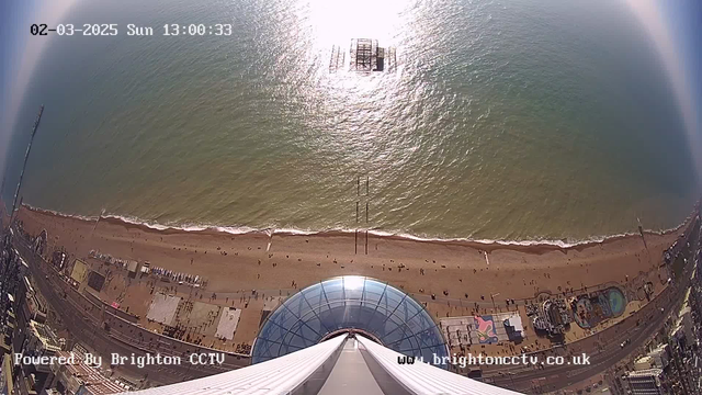 An aerial view captured from a webcam showing a beach scene. The perspective is from a high point, looking down at wide sandy beach with numerous people walking along the shore. The ocean water appears calm with gentle waves, reflecting sunlight. In the distance, there are remnants of a pier extending into the sea. The foreground features an architectural structure with a glass dome, likely part of an observation tower. Below, amusement attractions and various buildings can be seen along the beachfront. The image is timestamped with the date and time in the upper left corner.