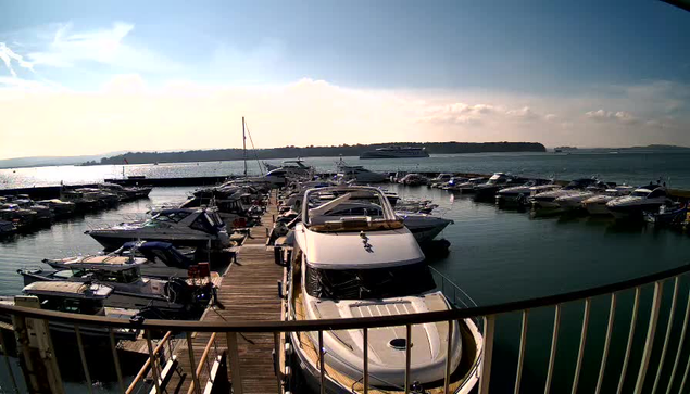 A marina scene with numerous boats docked at the pier, mostly yachts of various sizes. The water is calm and reflects the sky, which is partly cloudy with a few scattered clouds. In the background, a distant landmass can be seen across the water, and there is a larger vessel further out at sea. Sunlight illuminates the area, creating a bright and serene atmosphere.