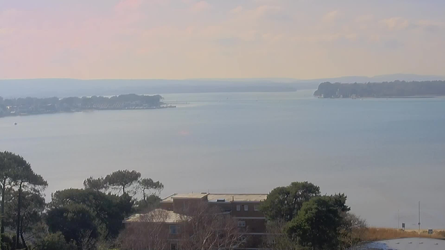 A calm body of water stretches into the distance, reflecting soft hues of blue and pink from the sky. In the foreground, there are trees with green leaves and a brown building with a flat roof. On the far side of the water, a shoreline is visible with buildings and boats scattered along the coast, edged by a gentle range of hills in the background. The scene conveys a tranquil atmosphere with a clear view of nature and architecture harmonizing.