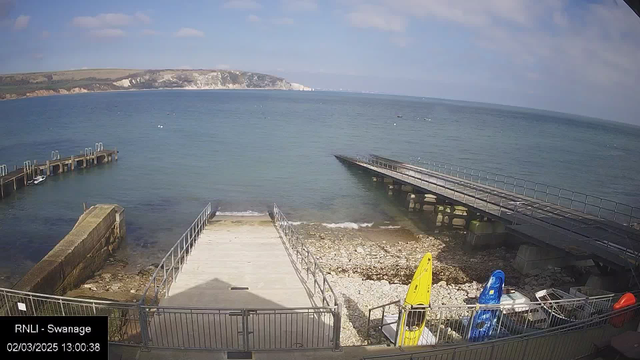 A coastal scene featuring a calm sea under a partly cloudy sky. In the foreground, there is a wooden dock extending into the water, with another dock visible in the background. To the right are two kayaks, one yellow and one blue, resting on a rocky shore. The shoreline has some large, smooth stones visible, and the water is clear with small waves lapping at the edge. In the distance, cliffs rise along the shore, contrasting with the blue of the sea and sky.