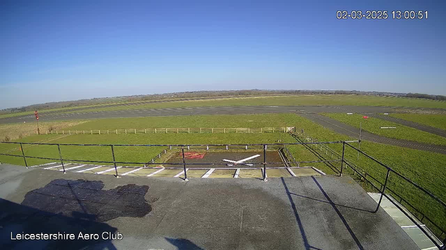 A clear blue sky is visible above an expansive green field. In the foreground, there is a rooftop area with a railing, where a section of gray surface is seen. A helipad with a white cross marking is located on the flat surface below, next to a small red area. In the background, the field extends to an airstrip with a few straight paths visible on the ground, while a fence runs along the edge of the grass.