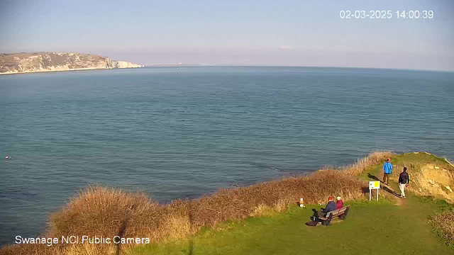 A coastal view shows calm blue waters extending out to the horizon under a clear sky. In the foreground, a grassy area is adorned with sparse bushes, and a wooden bench is occupied by two people. To the right, a man in a blue jacket walks along a path leading toward the cliff's edge. The cliffs are visible in the distance, displaying rocky textures and light-colored formations.