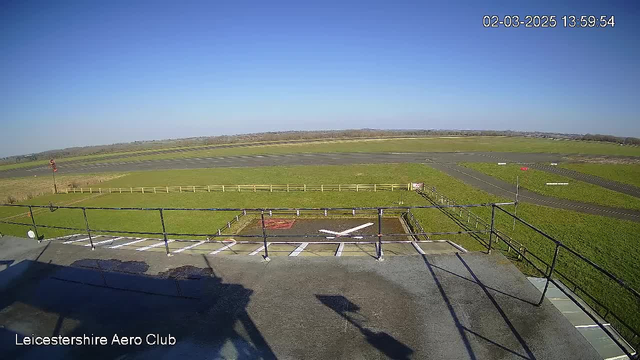 A clear blue sky fills the upper portion of the image, while a green grassy field occupies much of the foreground, bordered by a wooden fence. The image shows a section of an airstrip, with multiple taxiways visible. In the bottom left corner, there is a helipad marked with a white "X." The scene is well-lit, suggesting a sunny day, and there are no clouds in the sky.