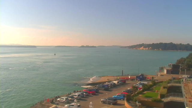 Image depicting a coastal scene from a webcam. The foreground features a parking area filled with various cars, predominantly red and blue, along with a path leading to the water's edge. In the background, the calm sea stretches towards the horizon with several small islands visible. The sky is clear and blue, suggesting a sunny day, with soft light reflecting off the water surface. On the right side, there's a building and landscaped area with greenery.