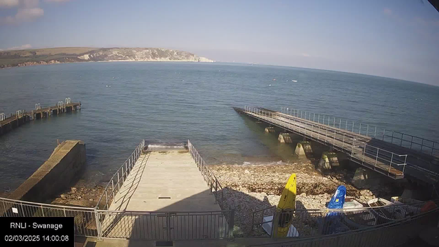 A view of the shoreline at Swanage, showing calm waters under a blue sky with some clouds. On the left, there is a wooden dock extending into the water, and to the right, a narrow ramp leads from a rocky beach to a flat surface. Two brightly colored kayaks, one yellow and one blue, are resting on the beach. The water is clear, with a few small waves gently lapping against the shore. In the background, white chalk cliffs rise along the coastline.