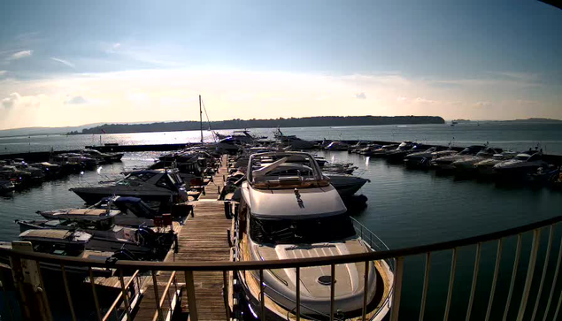 A view of a marina with numerous boats docked in calm water. The foreground includes a large white yacht at the end of a wooden pier. Several smaller boats are visible, with some moored closely together. The background features the horizon with hills in the distance under a bright blue sky, and sunlight reflecting off the water, creating a shimmering effect.