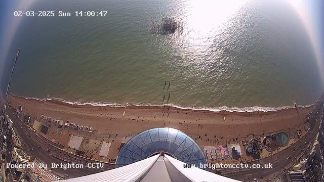 Aerial view from a high vantage point looking down at a beach with golden sand, gentle waves lapping at the shore. People are walking along the beach and there are colorful attractions and buildings visible. A pier is seen extending into the water, and there is a partially submerged structure in the ocean. The sky is clear with no clouds, and sunlight reflects off the water's surface, creating a glistening effect.