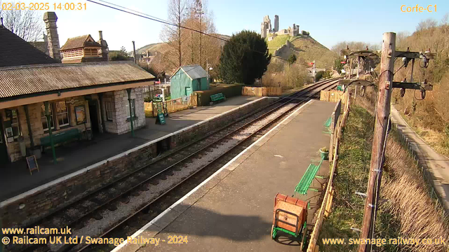 A view of Corfe Castle railway station on a sunny day. The foreground shows railway tracks with a wooden platform to the left. There are several green benches along the platform and a wooden cart on the right. Behind the station, there is a green shed and a fence. In the background, Corfe Castle stands on a hilltop, surrounded by trees and distant hills under a clear blue sky.