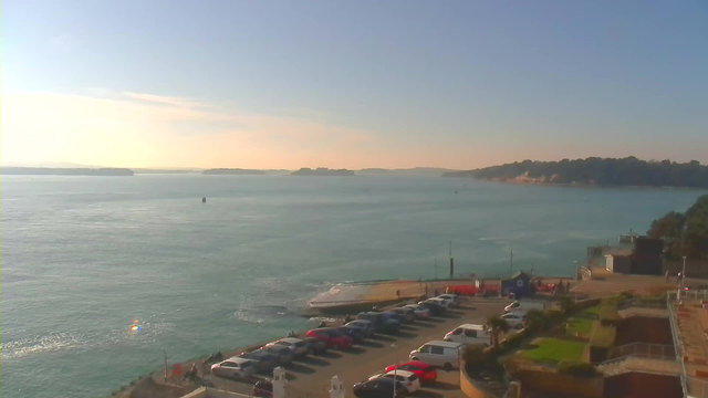 A coastal view featuring a calm blue sea under a bright sky with soft clouds. There is a line of parked cars along the shore, with some people visible near the water. In the distance, there are small islands and hills. The sun is shining, creating a reflective sparkle on the water's surface. On the right side, there are lawns and pathways leading to a building, with mountains in the backdrop.