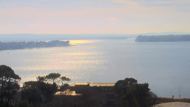 A serene view of a calm body of water reflecting soft pastel colors in the sky, with gentle ripples creating sparkles on the surface. In the distance, boats can be seen near the shoreline, framed by trees. A small colorful sailboat is navigating across the water, adding a vibrant touch to the scene. The background features a silhouette of land with trees and buildings, merging into a hazy horizon.