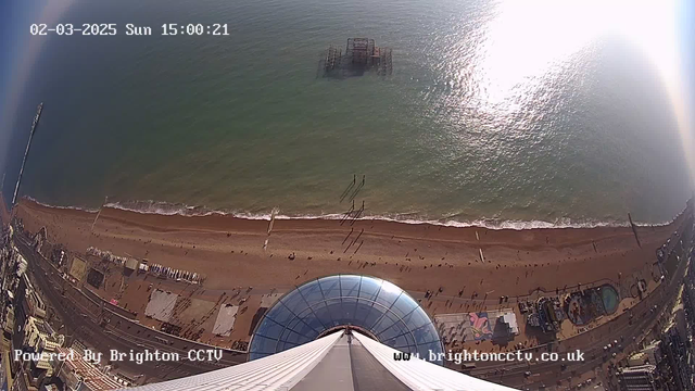 Aerial view of a beach with golden sand and gentle waves lapping at the shore. People are scattered along the beach and the water's edge. In the background, an old, rusted pier is visible partially submerged in the water. The scene is bright and sunny, with a large glass dome structure in the foreground. The horizon meets a calm sea, reflecting sunlight.