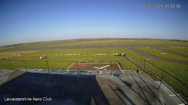 A clear sky is visible with bright blue colors. In the foreground, there is a flat rooftop with a railing and shadows. Below, an airfield covers a green expanse of grass, with a runway marked by black pavement. A white "X" shape can be seen on the ground, indicating a landing spot. In the distance, there are a few people gathered near a fence along the edge of the airfield, and some flags are visible. The scene is well-lit and captures a peaceful, outdoor setting.