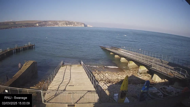 A seaside scene shows calm blue water and a rocky shoreline. There are two piers extending into the water, one concrete and one wooden. A few people are visible walking near the water's edge. In the foreground, there are kayaks stored on the ground, one yellow and one blue. The background features cliffs and green hills under a clear blue sky.