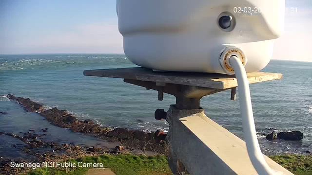 A white cylindrical object is mounted on a metal structure overlooking the sea. Below, a rocky shoreline is visible, with waves crashing against the rocks. The ocean appears choppy, and there is green grass in the foreground. The sky is clear with a few clouds, and the sun is shining brightly.