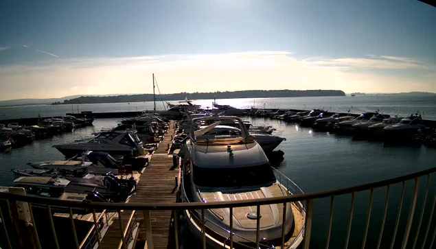 A scenic view of a marina under a bright blue sky with soft clouds. Numerous boats are docked in the marina, some tied to wooden piers, while others float on calm water. The sun reflects off the water's surface, creating a shimmering effect. In the background, a green hillside is visible across the bay. The image captures a tranquil and sunny day at the waterfront.