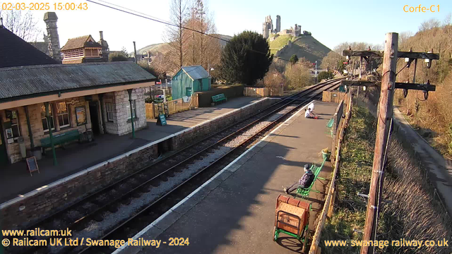 A sunny day at a quiet railway station. Two green benches are placed along a platform that runs beside two parallel train tracks. In the background, a stone building with a sloped roof is visible, along with a small wooden shed painted in teal. A prominent castle ruins atop a hill can be seen in the distance, while a few trees and a low fence are present nearby. On the platform, two people are seated, one appears to be engaged in an activity. The scene conveys a peaceful rural setting.