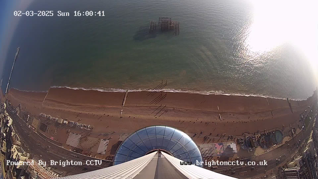 Aerial view from a high point overlooking Brighton beach, featuring a wide expanse of sandy shore and the blue ocean. The water is calm with gentle waves, and shadows cast by people walking along the beach are visible. In the distance, a partially submerged pier structure is detectable in the water. The foreground shows a modern structure with a curved glass roof. The time displayed in the top left corner reads "02-03-2025 Sun 16:00:41." The bottom of the image includes a watermark with the text "Powered By Brighton CCTV" and a web address.