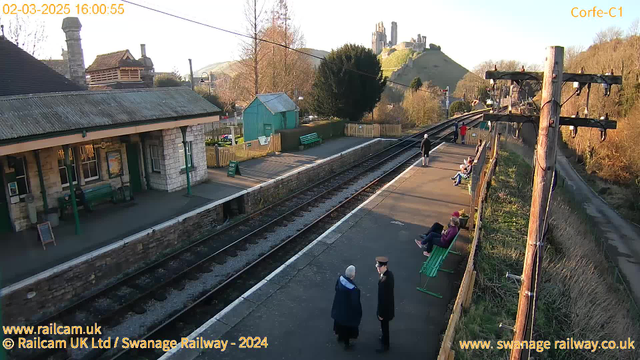 A view of Corfe Castle train station featuring a stone platform with several people sitting and standing. On the left, there's a small building with a green bench and a sign. In the background, Corfe Castle is visible on a hill. The scene is set in daylight, with trees lining the platform and a fence in the background. The railway tracks run alongside the platform.