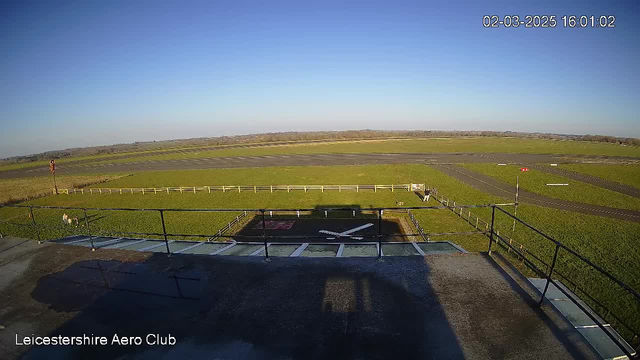 A clear blue sky stretches across the top of the image. Below, there is a grassy field with a runway in gray asphalt visible. In the foreground, a fence borders the grassy area, and there are markings on the ground that appear to indicate a helicopter landing zone. The scene is well-lit with sunlight, and the surrounding area is flat with trees visible in the distance. A small, red object can be seen on the right side of the runway. The bottom left corner displays the name "Leicestershire Aero Club."