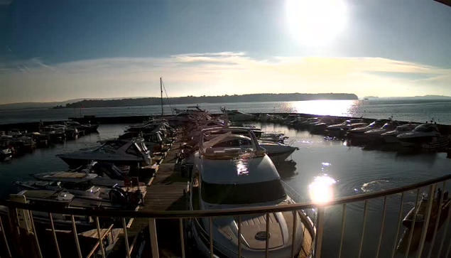 A marina scene with numerous boats docked in the water on a sunny day. The sun is shining brightly in the sky, casting reflections on the water's surface. In the background, there are hills or land masses. The wooden docks extend along the edge of the water, and some boats are clearly visible, while others are partially hidden. The overall atmosphere is tranquil and bright.