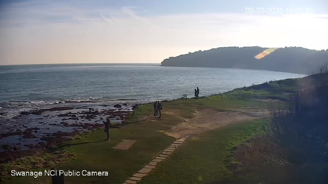 A coastal scene with a calm sea under a clear sky. In the foreground, a grassy area with a pathway made of stones leads towards the shore. Several people are seen walking along the path and near the water's edge, while others are standing, enjoying the view. In the background, a green hill rises above the sea, and the sun appears low in the sky, creating a warm glow. The image captures a peaceful, serene atmosphere.