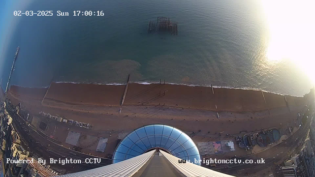 A high-angle view of a beach scene taken from a webcam. The image shows a sandy beach with gentle waves lapping along the shore. A pier extends into the water from the left side, with a ruined structure visible partially submerged a bit further out. In the foreground, there is a circular glass structure, possibly the top of a tower or observation point, with visible rooftops and roads. The sky is mostly clear with some soft lighting reflecting off the ocean surface, indicating it's late afternoon. The date and time in the top corner reads "02-03-2025 Sun 17:00:16." The bottom corner displays "Powered By Brighton CCTV" and a website link.