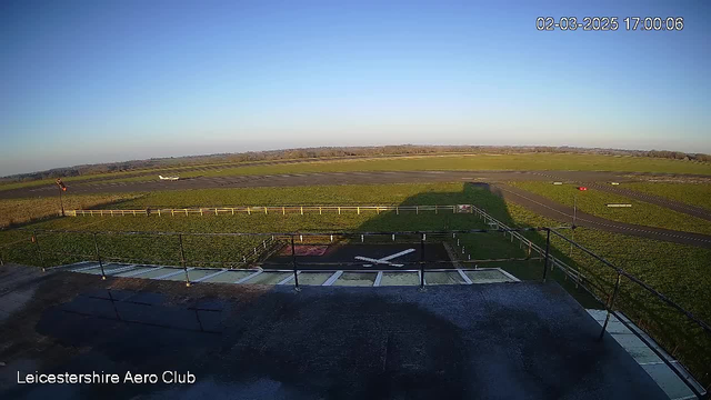 A clear blue sky stretches over an open landscape with green grass and a runway in the foreground. The runway is marked with lines and has a small white aircraft positioned on it. In the lower left corner, a fenced area with a helipad symbol can be seen. The scene is tranquil, with no visible people, depicting the remote view from the Leicestershire Aero Club.