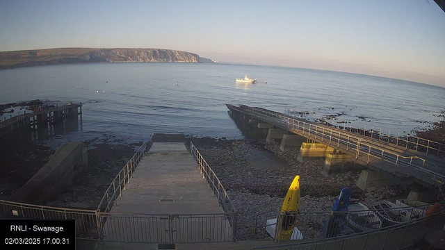 A serene coastal scene at Swanage, featuring a calm water surface reflecting the blue sky. In the foreground, a stone ramp leads down to the water, bordered by a metal railing. To the right, several colourful kayaks are parked on the rocky shore, with a bright yellow kayak prominently displayed. In the distance, a small fishing boat is anchored, while natural cliffs rise along the shoreline in the background. The scene is bathed in soft, warm light, indicating early evening.