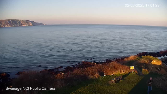 A coastal scene showing calm water reflecting soft light, with a rocky shoreline on the left and green grass in the foreground. In the lower right, a person stands near a bench, facing the water. There are sparse signs and some vegetation along the edges. The sky transitions from light blue to soft pink near the horizon.