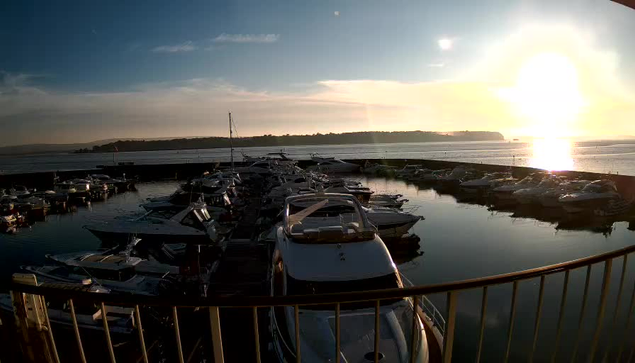 A scenic waterfront view featuring numerous boats docked in a marina. The sun is setting on the horizon, casting a warm golden glow over the water. Reflections of the boats can be seen on the calm surface of the water, with a backdrop of distant hills and a clear sky. The scene is peaceful, depicting a tranquil evening by the harbor.