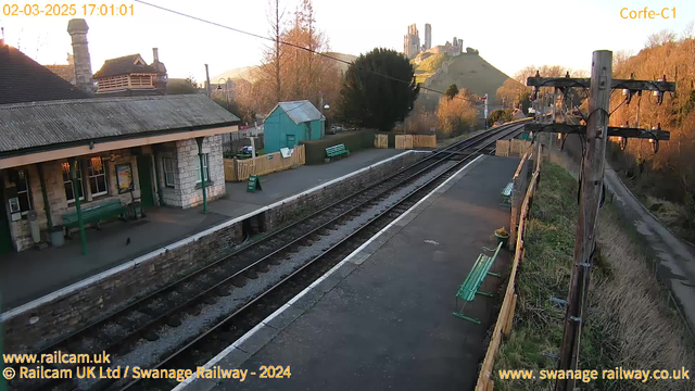 A view of a train station in Corfe, with stone buildings featuring a sloped roof and green benches on the platform. The railway tracks run into the distance, leading to a hillside with ruins in the background. The scene is illuminated by a warm sunset, casting long shadows. There is a sign that reads "WAY OUT" near a wooden fence, and a green building is visible in the background among trees.