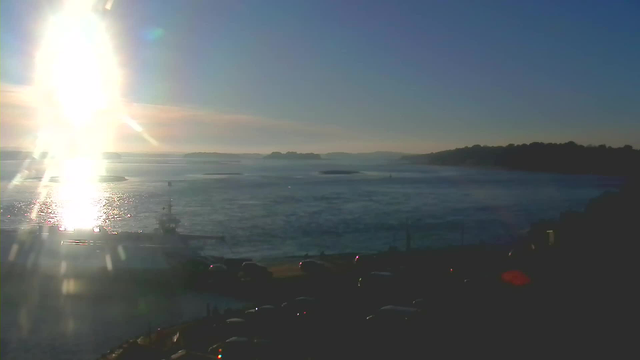 A bright sun reflects on the water of a bay, with a clear sky in the background. The horizon is lined with distant land, and there are small boats on the water. In the foreground, a road is visible with several parked cars. The scene captures a serene and sunny coastal landscape.