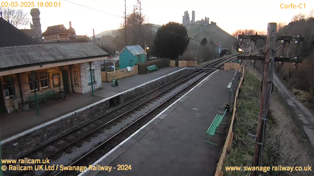 The image shows a view of Corfe Castle railway station during sunset. The foreground features a platform with a few green benches and a stone building with large windows, possibly a ticket office. Beyond the platform, two sets of railway tracks run parallel, leading off into the distance. To the right, there is a wooden fence enclosing the station area. In the background, the silhouette of Corfe Castle is visible on a hill, with a gradient sky transitioning from light orange to a deeper blue indicating evening. A wooden utility pole with wires stands to the right, and there are trees in the surrounding area.