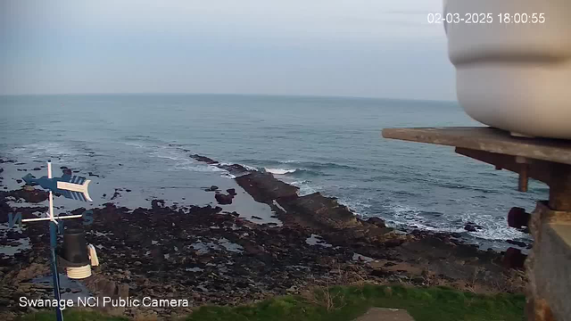 A coastal scene captured by a webcam shows the ocean with gentle waves lapping against rocky shores. In the foreground, there is a white and blue weather vane shaped like a fish, indicating wind direction. To the right, the camera captures part of a white container, possibly related to the weather station. The sky is overcast and light gray, while the water appears calm with varying shades of blue. The date and time stamp in the corner reads "02-03-2025 18:00:55."