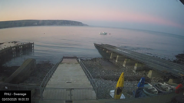 A serene seaside scene at dusk, depicting calm waters with a small white boat floating in the distance. The foreground shows a rugged shoreline with scattered rocks and a concrete ramp leading down to the water. A metal railing runs alongside the ramp, and brightly colored kayaks—yellow and blue—are positioned on the side. The sky transitions from pale pink to soft blue as evening approaches, with a few buoys visible on the water's surface.