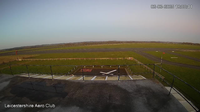 A wide view of an open grass field and a small airstrip. The foreground shows a black rooftop edge with a white emblem resembling an airplane in the center. In the background, a green field stretches to the horizon, bordered by a wooden fence. A tall red wind direction indicator is visible to the left, while the airstrip has marked lanes and areas for movement. The sky is clear with a pale blue hue, indicating early evening light. A timestamp in the corner reads 18:00:41 on March 2, 2025.