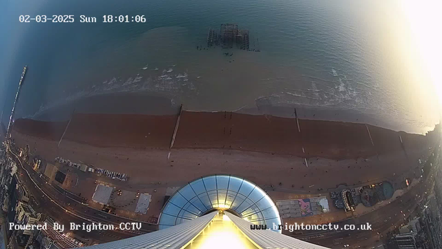 A view from above a coastline with sandy beach and waves. The image captures the shore, where people can be seen walking along the beach. A pier structure extends into the sea. The sky is partially clear, indicating a possible sunset with soft lighting. The bottom part shows the base of a tall building with a circular glass design. The date and time are displayed in the top left corner.